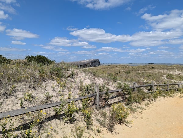 A wooden fence snaking through low dunes with scrubby vegetation, and a causeway rising up in the distance