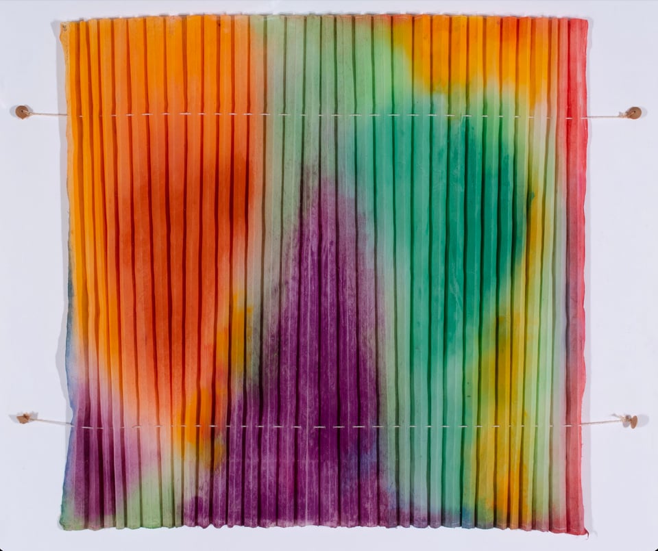 A square of pleated paper covered with abstract, organic shapes in orange, green, purple, pink, and yellow with blurred edges. The pleats are held in place by two pieces of string running across the top and bottom of the piece and affixed to the wall.