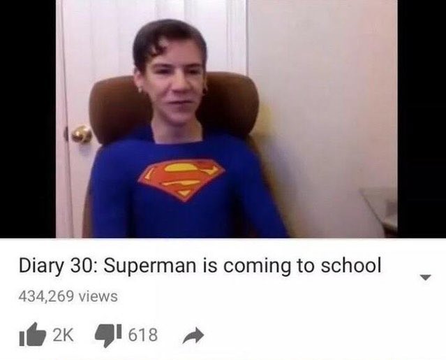 A screenshot of Dax Flame's vlog entry "Superman is coming to school". A skinny teenage boy with a light skin tone and brown hair is dressed in a Superman costume, with his hair slicked over and what look like hoop earrings dangling from his ears. He is smiling at the camera.