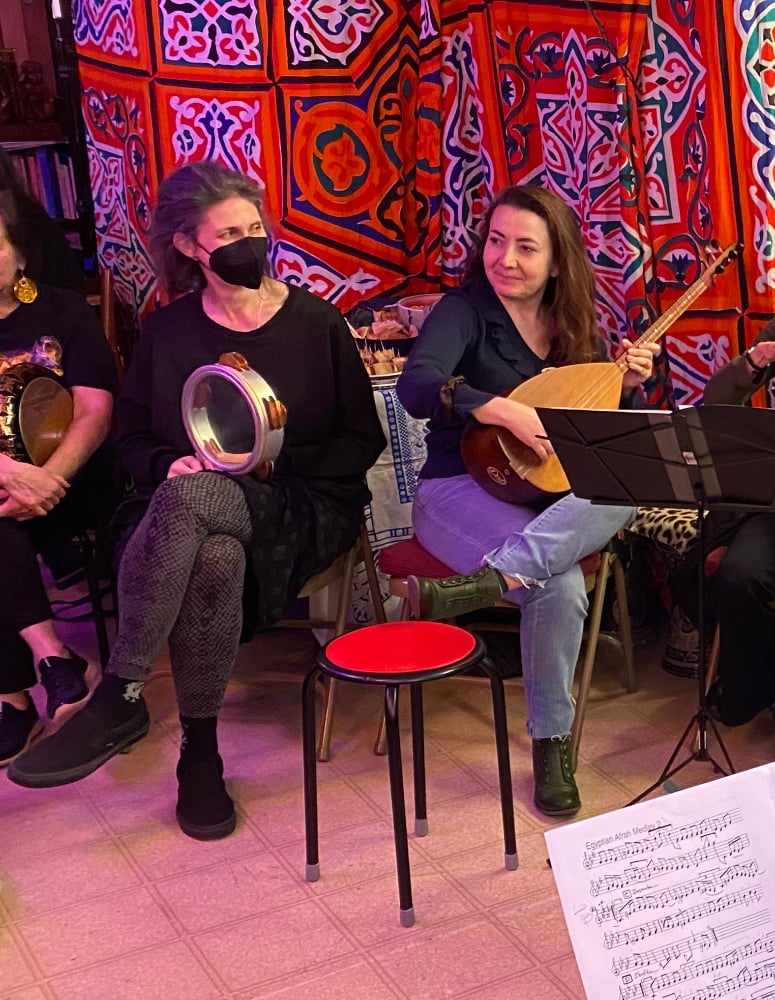 Two people sit in a colorful studio, one with a riq the other with a baglama