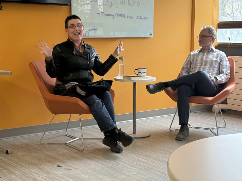 Ada Hoffmann is wearing short hair, glasses, a leather jacket, jeans, and black running shoes. They are sitting in a red chair in front of a yellow wall, waving their hands in an expressive gesture. They look happy and excited. Beside them is a small table with a water bottle, copy cup, and book. On the other side of the table a man with gray hair sits. He is wearing a striped shirt, jeans, and brown shoes. He is the event host.