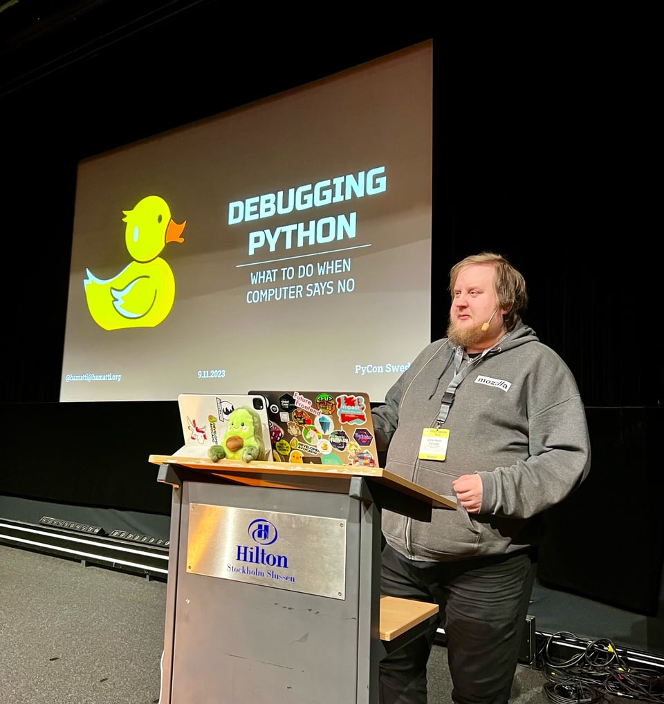 Juhis standing on a stage with a laptop in front of him on a speaker podium. Behind him, on a large screen, a yellow rubber duck illustration with the text Debugging Python - What to do when computer says no
