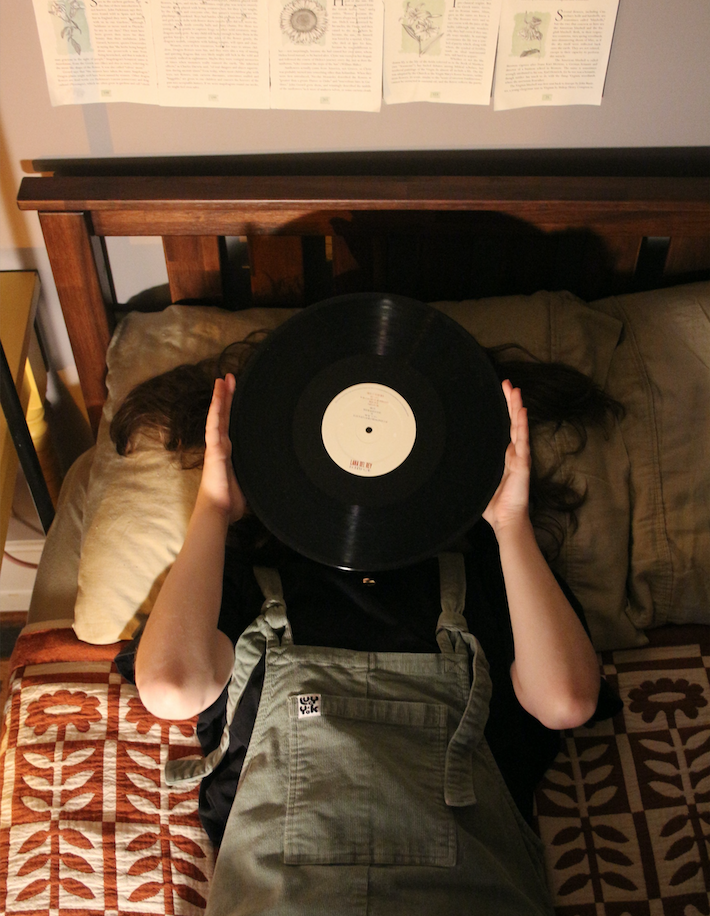 Copeland is lying down on their bed, holding a vinyl record over their face. They're wearing green corduroy overalls over a black T-shirt, and their bedspread has a cream and terracotta floral pattern on it.