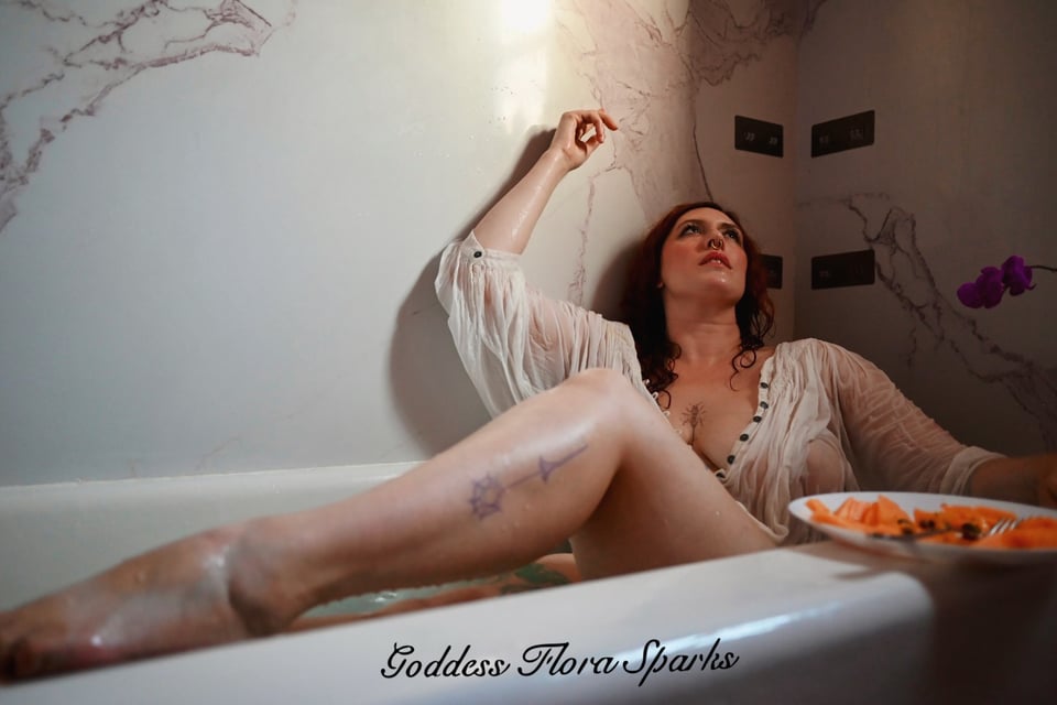 Flora Sparks is posing dramatically in her bathtub. She is wearing a white shirt that is soaking wet and draping beautifully. Her leg is up on the side of the bathtub and poses with pointed toes