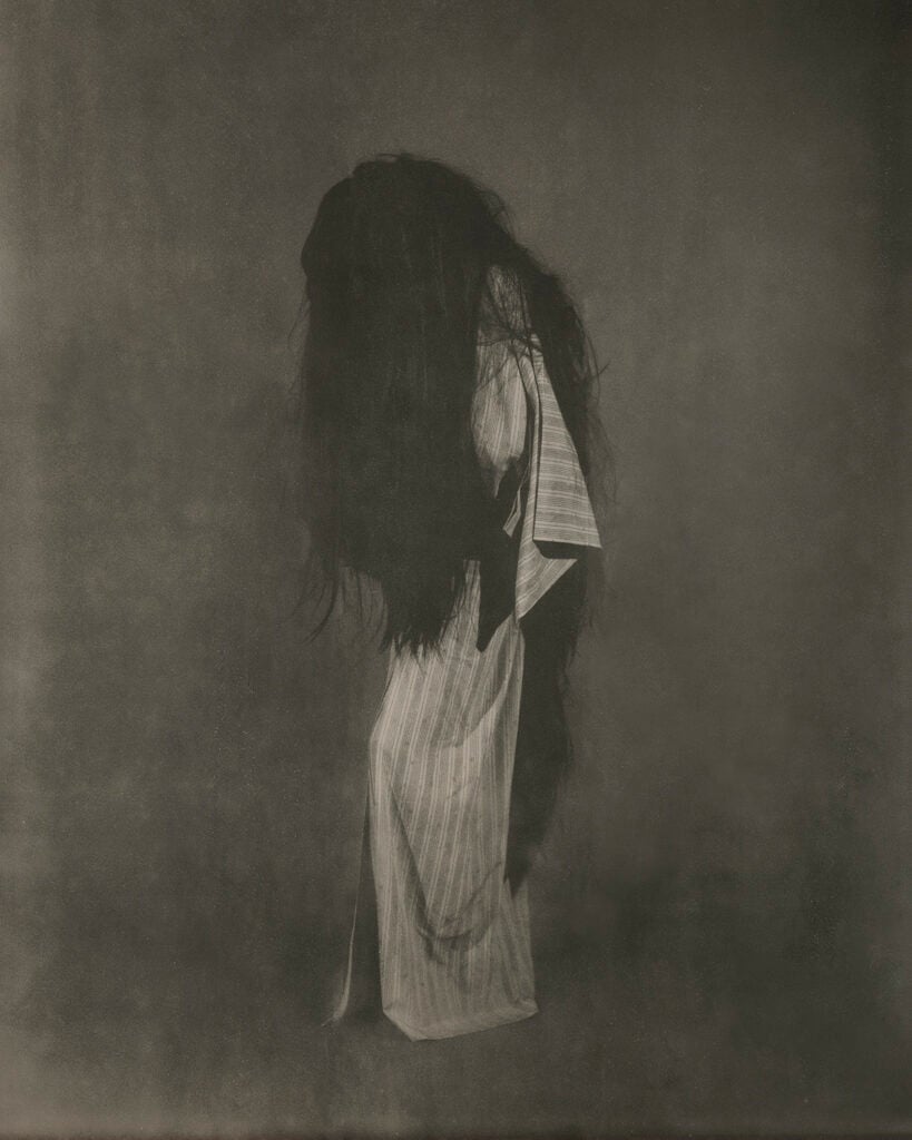 A haunting sepia-toned photograph of a figure with extremely long, dark, wild hair cascading down and obscuring their face and upper body, wearing a pale vertically striped garment, standing against a mottled grey-brown backdrop.