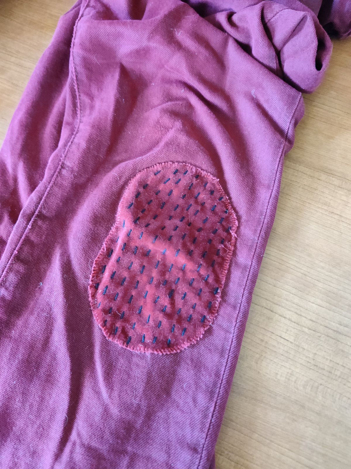 Photo of 1 leg of a pair of maroon-colored pants. On the knee of the pants is a velour patch of the same color with long black running stitches.