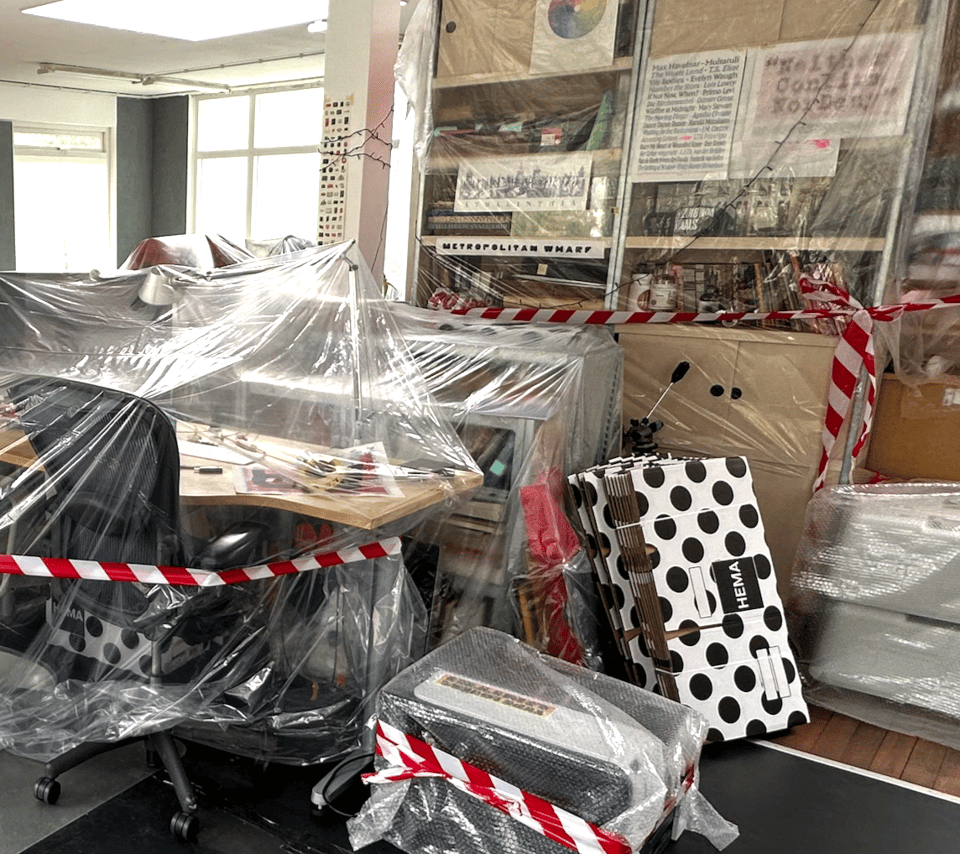 A photo of a desk, a printer and several bookshelves, covered in transparent plastic and wrapped in red and white tape.