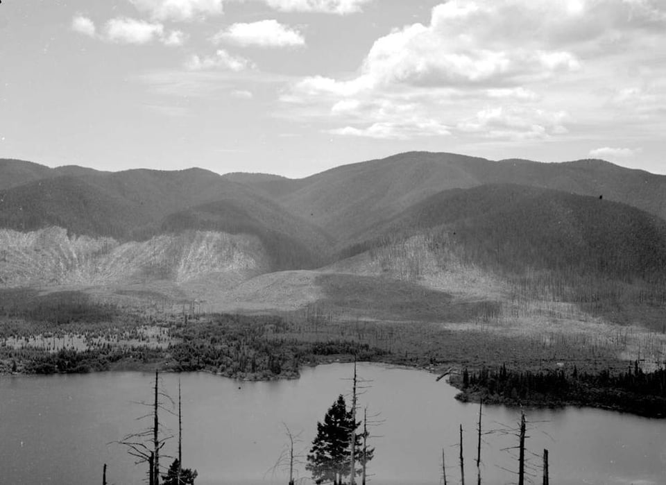 Black and white photo of mountains with water in the foreground. Some of the mountains are stripped of trees