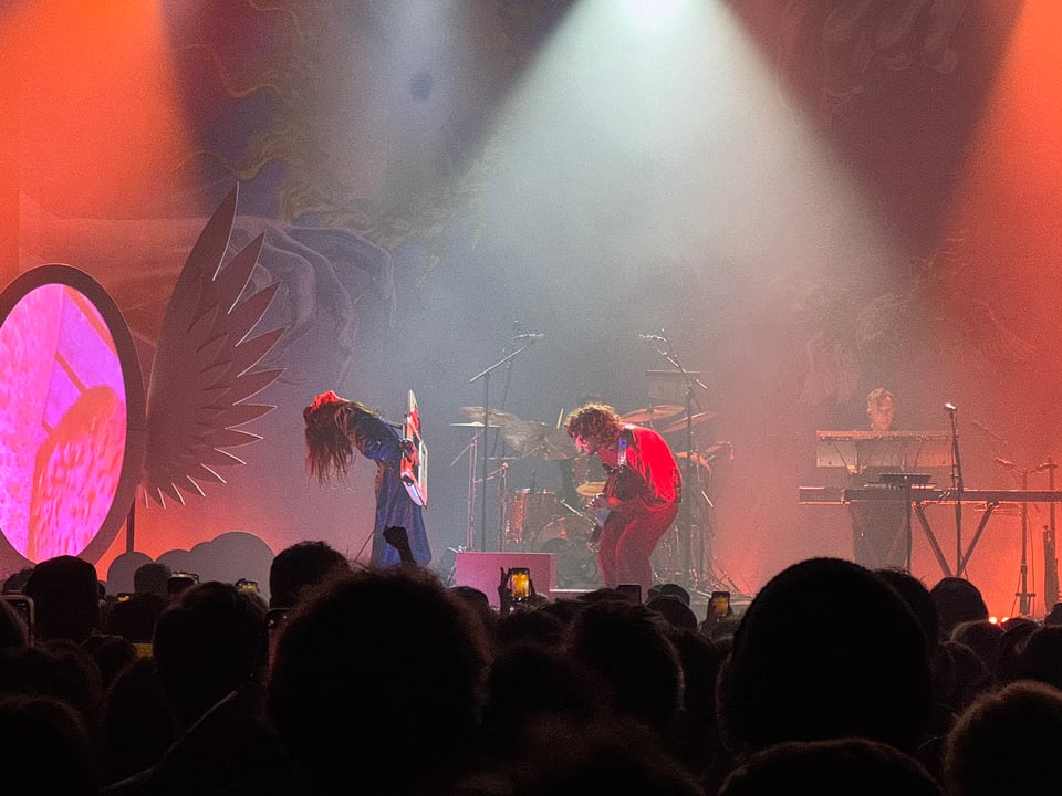 Magdalena Bay performs at Terminal 5. Mica and Matt stand facing each other playing a keytar and bass, respectively. A large mirror with wings and visuals inside it is visible on the left.