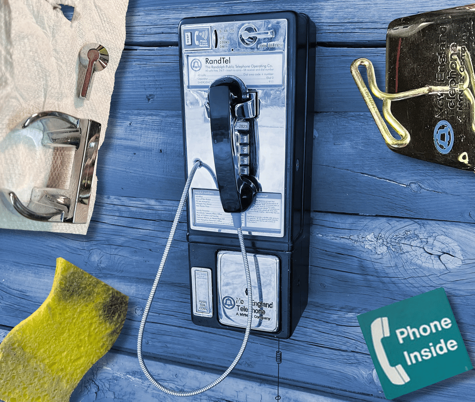 A payphone installed on a wall made of thick wooden beams. It looks like it's on the outdoors of an old fashioned wooden building. It has cut-out art around it, including a yellow scrub pad and a green sign saying "phone inside"