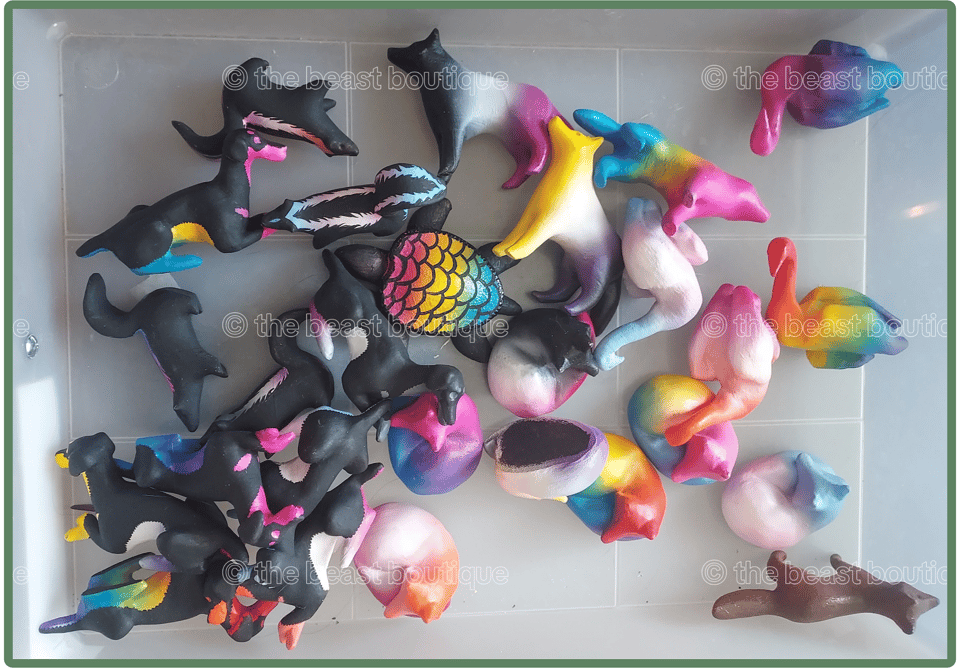 A photo of assorted figurines in a plastic drawer, including wolves, geese, foxes, and dobermanns, all painted in the colors of various pride flags.
the shop name, the beast boutique, watermarks the image.