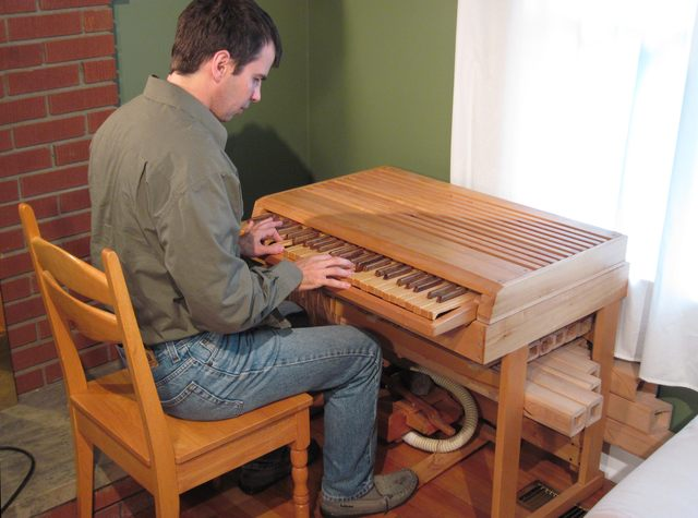 A man sitting at a small, brown wooden keyboard. He is sitting on a wooden chair and his hands are both playing the keys. He is dressed in jeans and all of green shirt. Beneath the Oregon, you can see several wooden square pipes.
