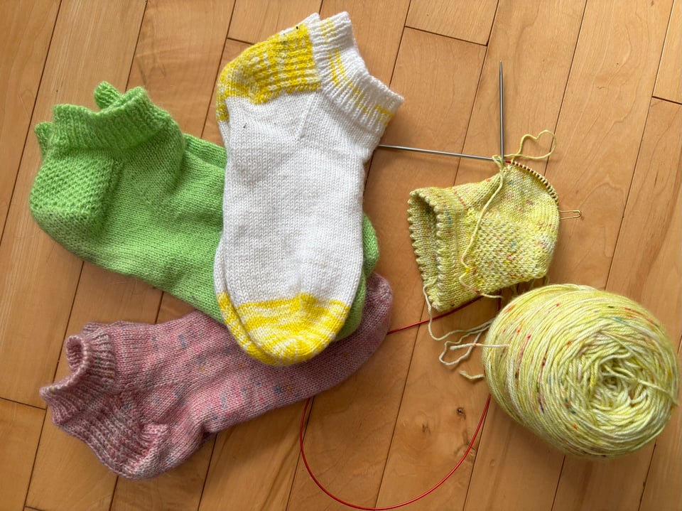 An array of ankle-length "shorties" handknit socks, clockwise from bottom left: pink with multicolour speckles, bright green with an eye-of-partridge pattern on the heel, white with lemon-yellow contrast, a work-in-progress on metal circular needles with a long red cord, next to its ball of yarn; the in-progress sock is pale yellow with multi-colour speckles and has an eye-of-partridge heel and a folded picot cuff.