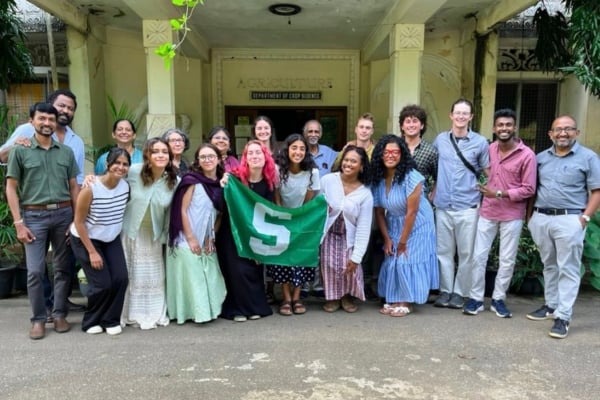 A group of students and faculty members are posing for a photo in front of the Agriculture Department building. They are standing together, with some holding a green flag featuring a white 'S'.