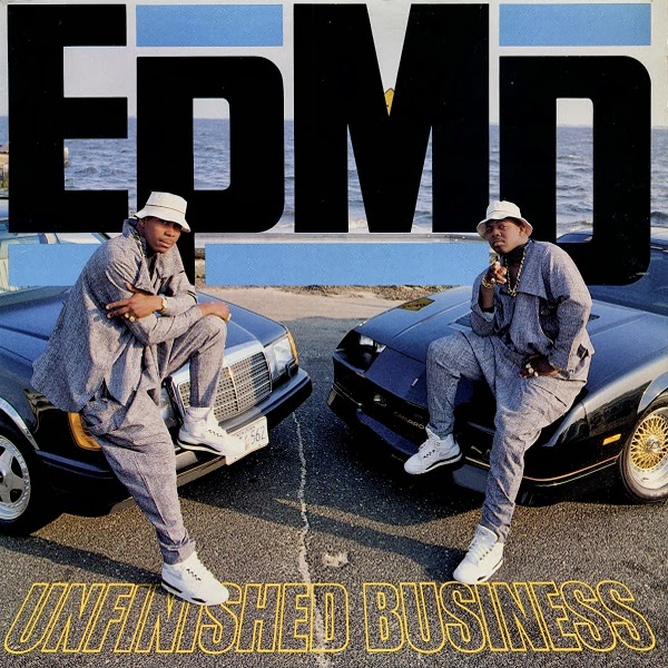 Two young Black men, dressed in identical loose-fitting grey clothing, white sneakers and white kangol hats, each sit on the hood of a car. They look at the camera. The cars are parked facing each other at an angle. Beyond them is the ocean.