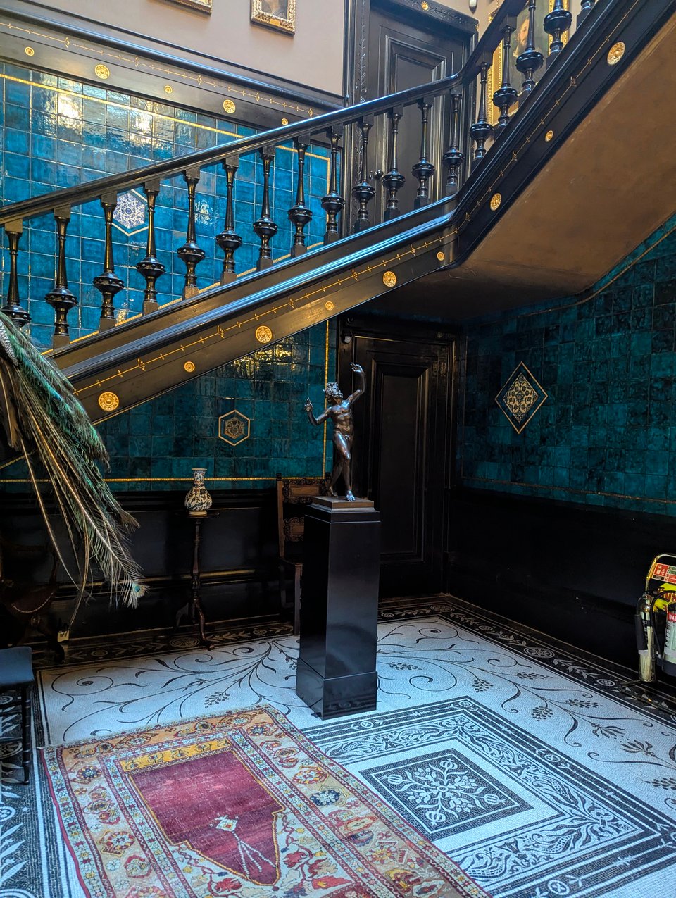 A photo of the bottom of an ornate set of black and gold stairs, the walls tiles in vivid blue, the floor a black and white mosaic with a small statue on a narrow plinth on the center.