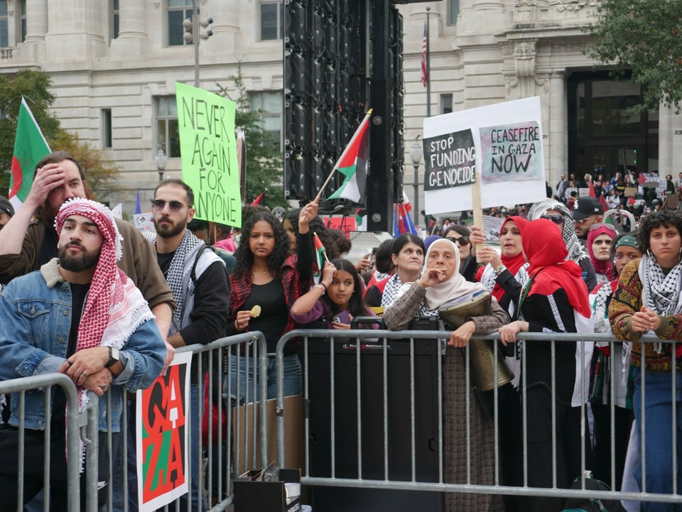 Protestors behind metal gates with a Gaza sign on it. Many are wearing keffiyehs and some are holding signs: Stop funding Genocide. Ceasefire in Gaza now. Never again for anyone.
