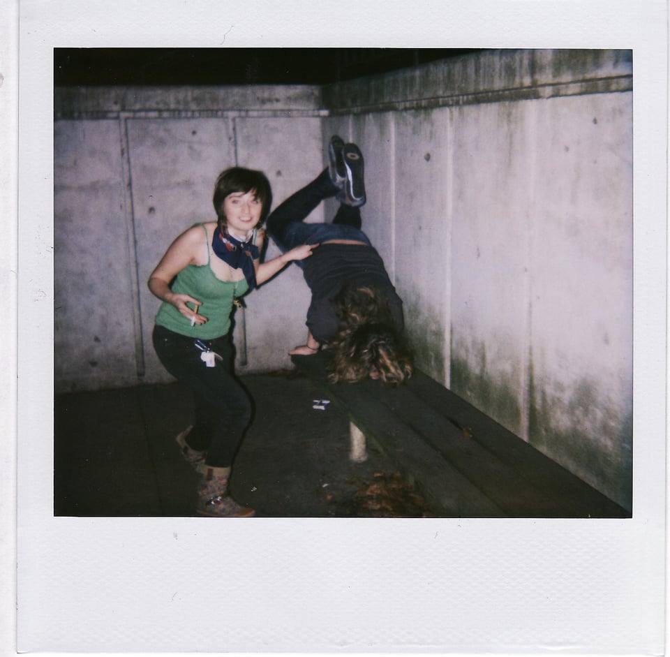 An 18-year-old Sandy smoking a cigarette in the smoking pit while her friend does a handstand on a bench in the background.