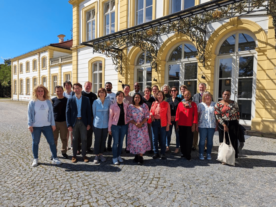 21 people of various dispositions wearing a variety of different colors in front of a light yellow and white palace front entrance. It is bright and sunny with clear skies.