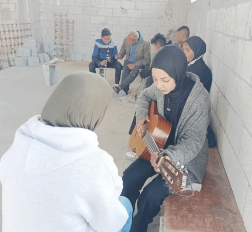 on a bench in a room made of cinderblocks, people seated and two people playing musical instruments 