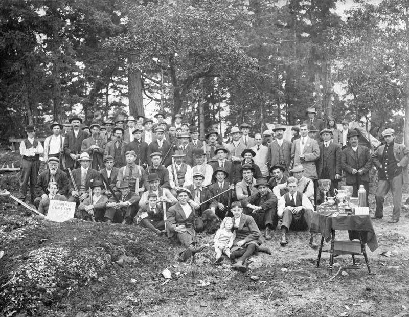 Black and white photo. A group of mostly men wearing hats holding guns and posing for photos. Off to the right of the photo is a table with a number of trophies