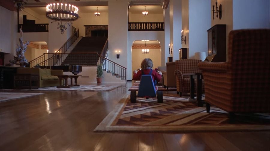 Danny riding in a low-slung tricycle around the Overlook Hotel. We see Danny’s back, and the rear of the bike. In the background is a large staircase, and on either side of Danny are pieces of the hotel’s furniture.