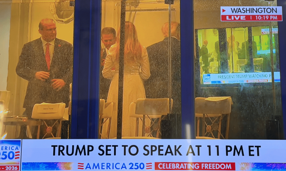 Photo of Fox News screen -- showing Donald And Melania Trump and a couple of men from a distance in a glass enclosure, ahead of Trump's July 4 speech in Washington. On the wall is a TV screen showing the same Fox news image and Trump is watching himself on the TV.