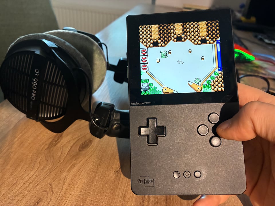 A picture of a handheld videogame device, running the game PachiPals, next to a pair of headphones on a table.