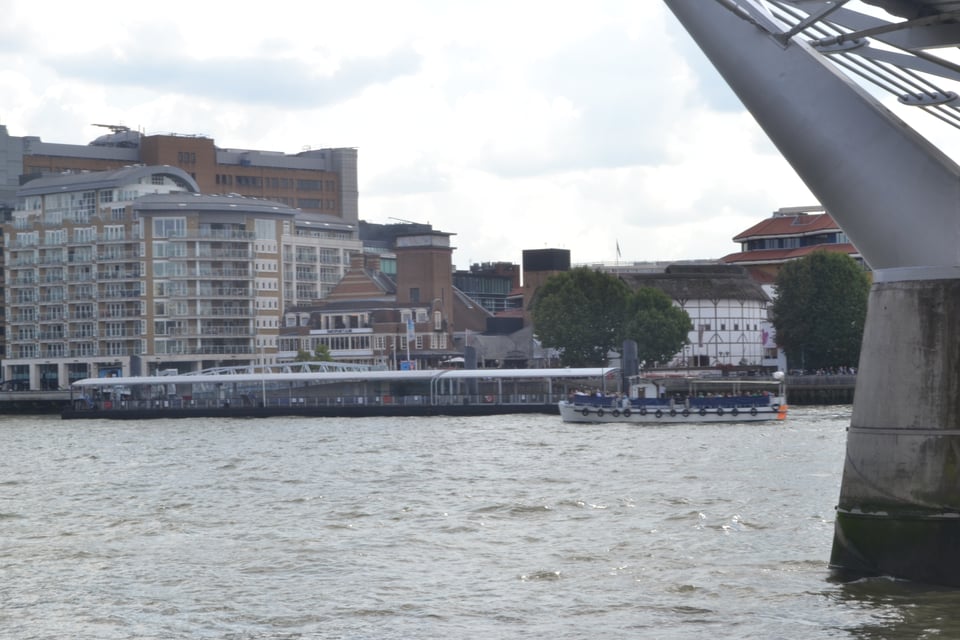A broad river seen from under a bridge, with a boat on the water, a dock, and large buildings on the other side, including an apartment building with balconies and a smaller half-timbered building.
