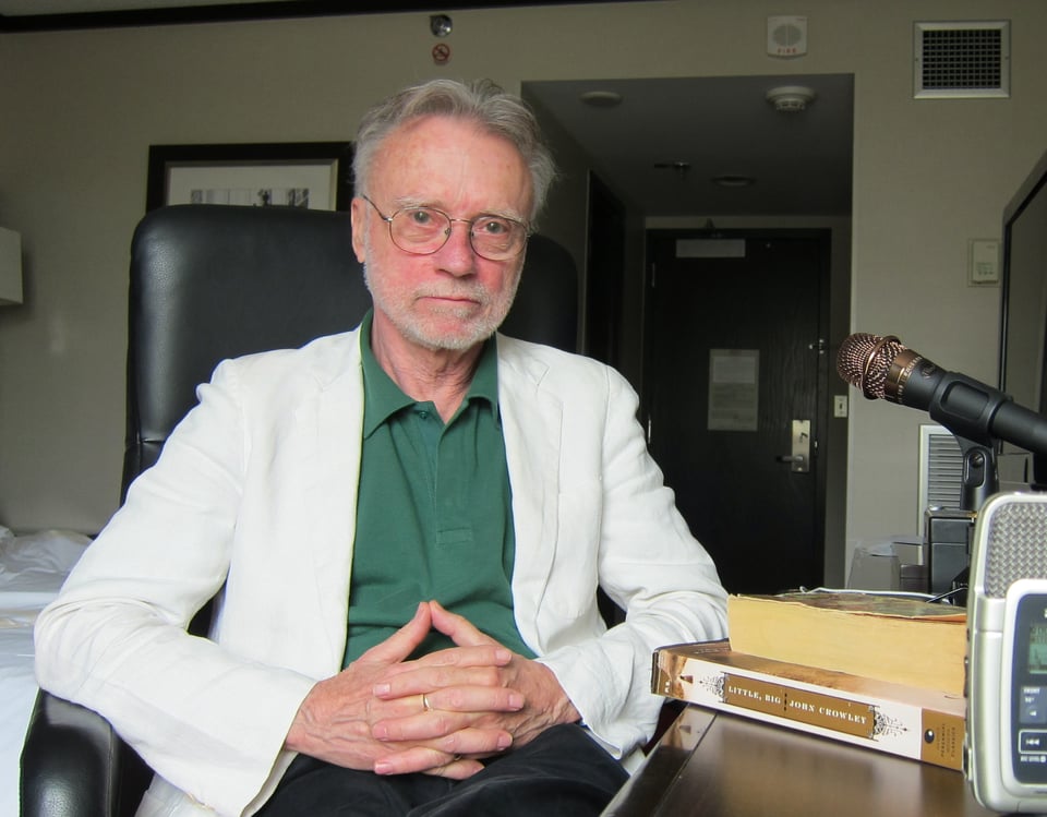 photo of author John Crowley in 2013, sitting at a table in a hotel room beside copies of his novel LITTLE, BIG, with a microphone on the right side of the show. He's wearing a white sport coat and green button-down shirt