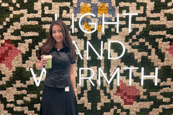 Photo shows JMC student Elena Sweeney in front of an artistic background with lights that read "Light and Warmth." She is wearing a black shirt and skirt and is holding a matcha latte.
