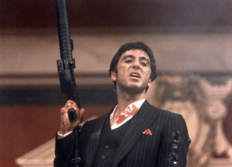 Movie still of Al Pacino, blood on his suit, holding up his high-powered machine gun and scowling at his enemies.