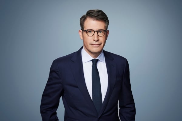 A man wearing a dark suit, white shirt, and tie stands against a plain gray background. He wears glasses and looks directly at the camera with a neutral expression.