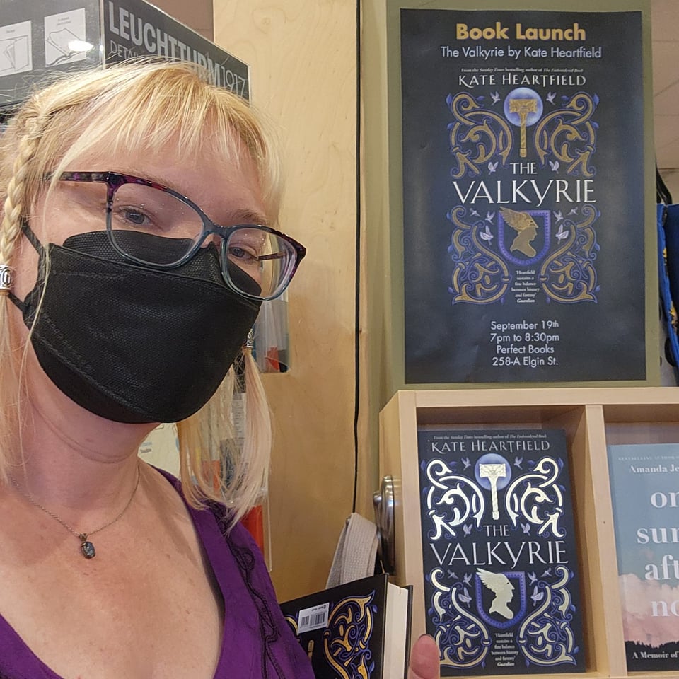 Kate is wearing a purple medieval style blouse, silver run beads in her hair, and a black mask. She is standing in front of a display shelf and poster for The Valkyrie.