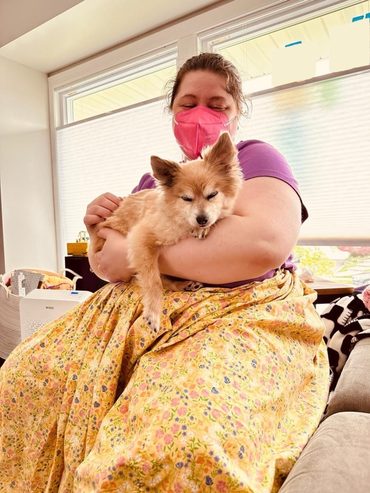 Photo of Jessi holding an older dog in their arms on a couch inside a home with bright windows and children's toys. The dog is small and fluffy and golden with white around his eyes and muzzle; his eyes are closed, and his front paw drapes over Jessi's arm. Jessi is a fat, white person with brown hair pulled back; they wear a purple t-shirt with a floral yellow skirt and a bright pink KN95 mask.