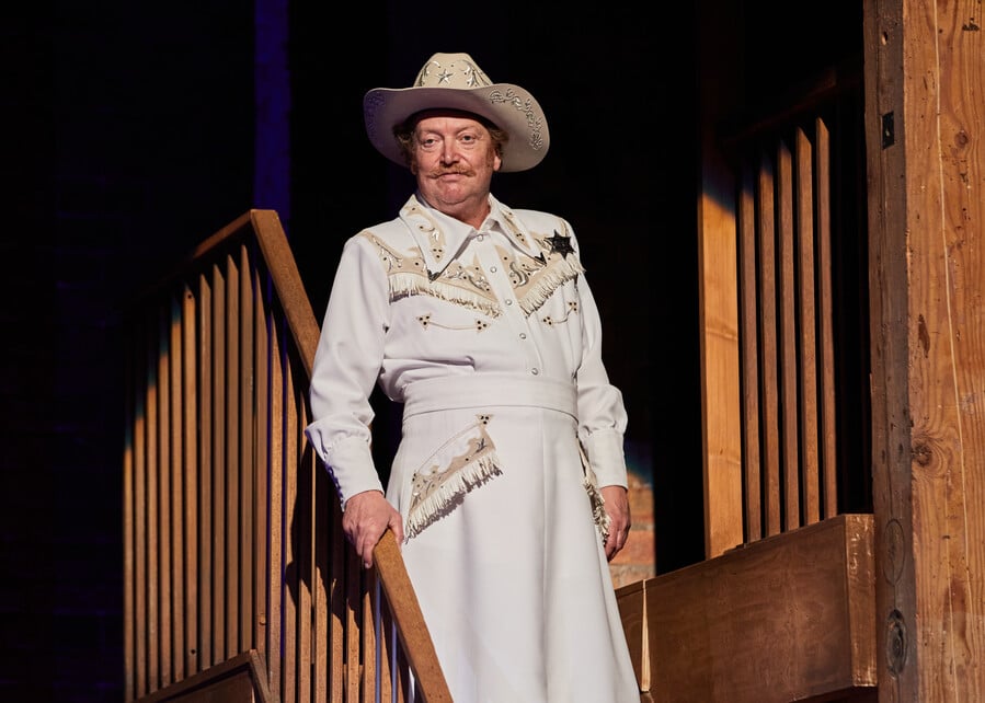 An older man standing in a spotlight with an all white and gold sheriff-themed dress and also a white cowboy hat.