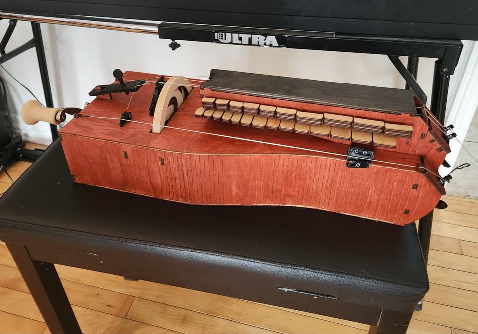An instrument lying on a piano bench and nearly the same length, stained cherrywood red, with a wheel and a crank and a keybox and four strings.