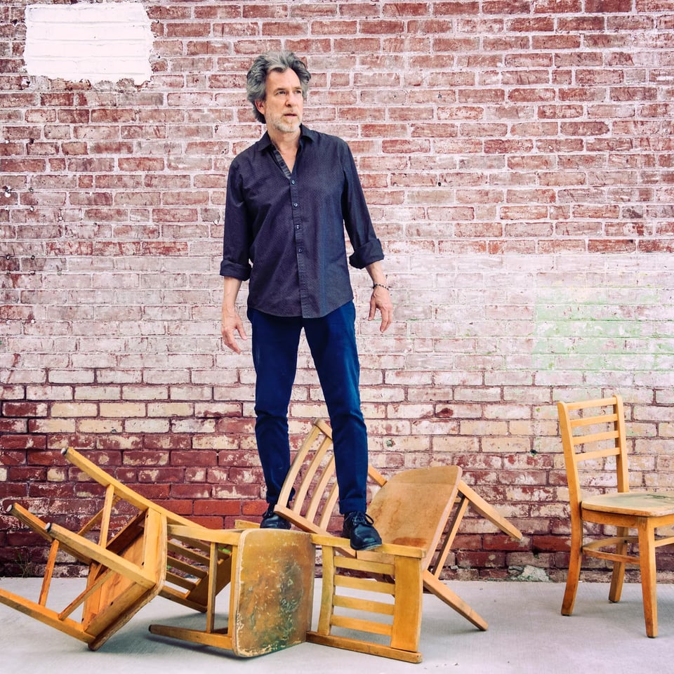 A man with a thatch of gray-brown hair wearing an untucked blue button-up shirt and blue chinos stands on an overturned chair in front of a brick wall.