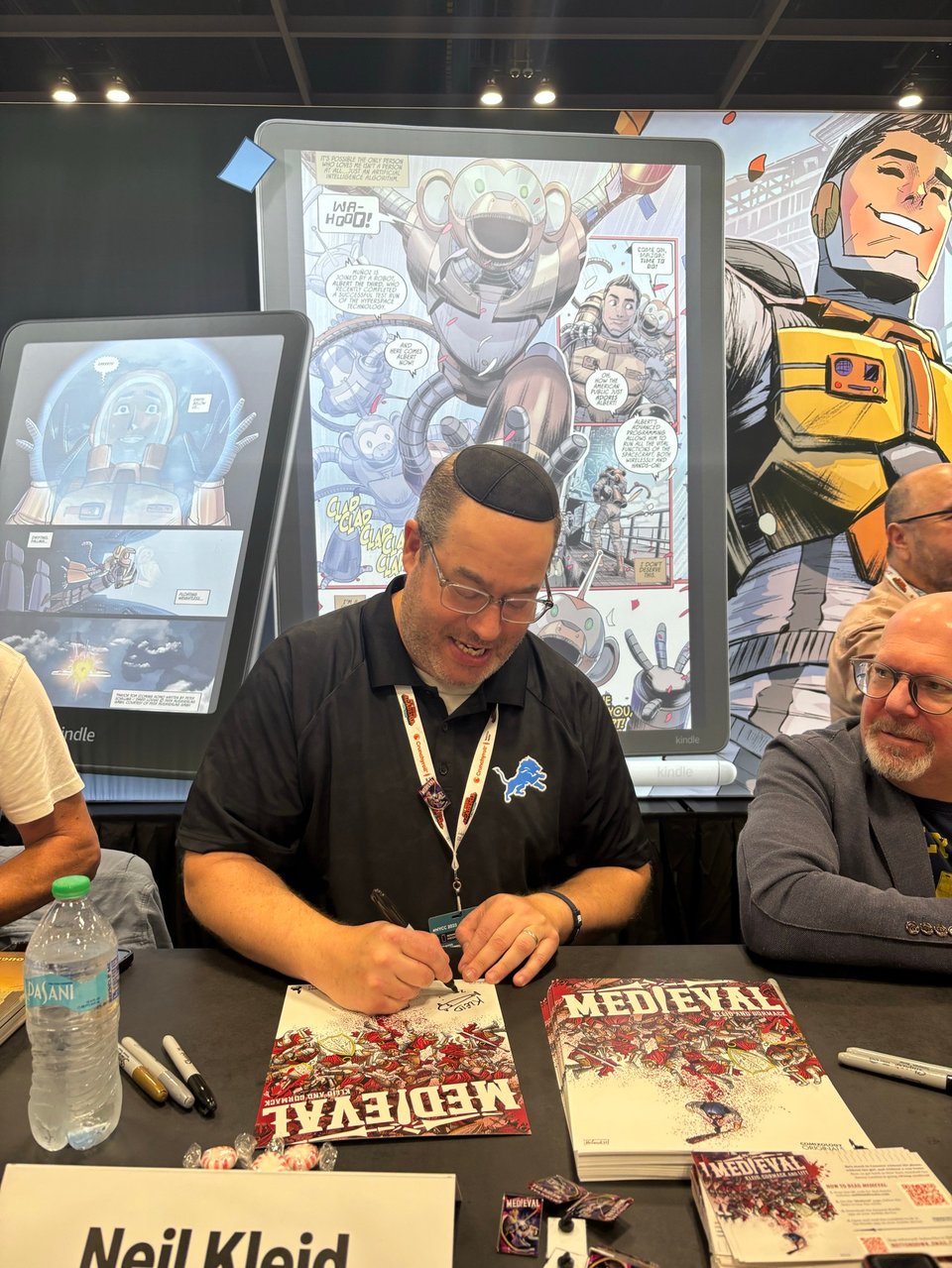 neil signing medieval posters at the comixology booth with marc guggenheim looking on