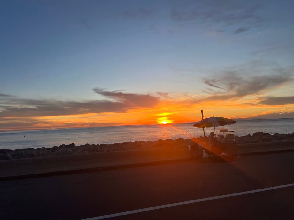 Sunset over the Pacific ocean. A person with a fruit cart and umbrella are in the foreground. The photo was taken from a moving vehicle.