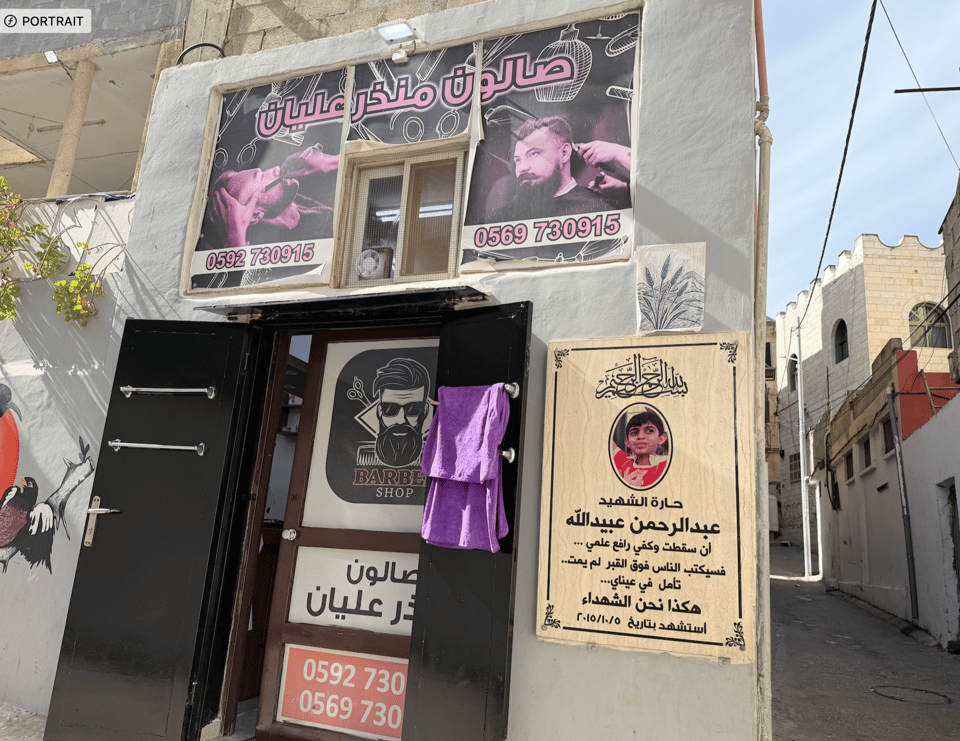 Photograph of a barber shop entrance with open black doors and a purple towel hanging on one door, featuring a sign above with a man getting a haircut and phone numbers with writing in Arabic. Next to the door is a memorial plaque with a photo of a young boy and Arabic text, set against a backdrop of a narrow street with many buildings
