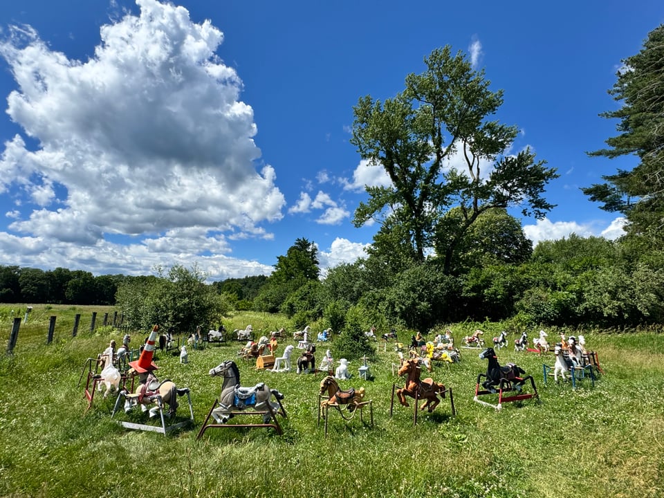 A large outdoor collection of vintage rocking horses of various colors and sizes arranged on a grassy field under a bright blue sky with fluffy white clouds.