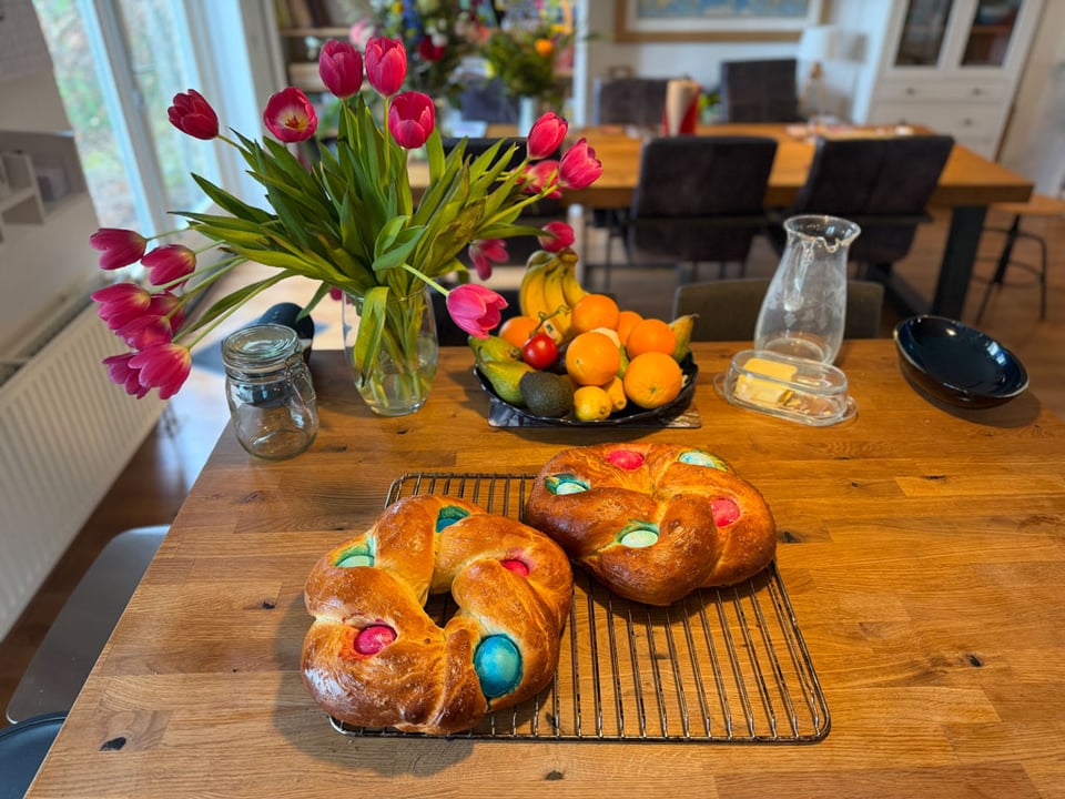Two braided loaves of Easter bread cool off on a rack on a kitchen island. A vase filled with drooping pink tulips is behind the bread.