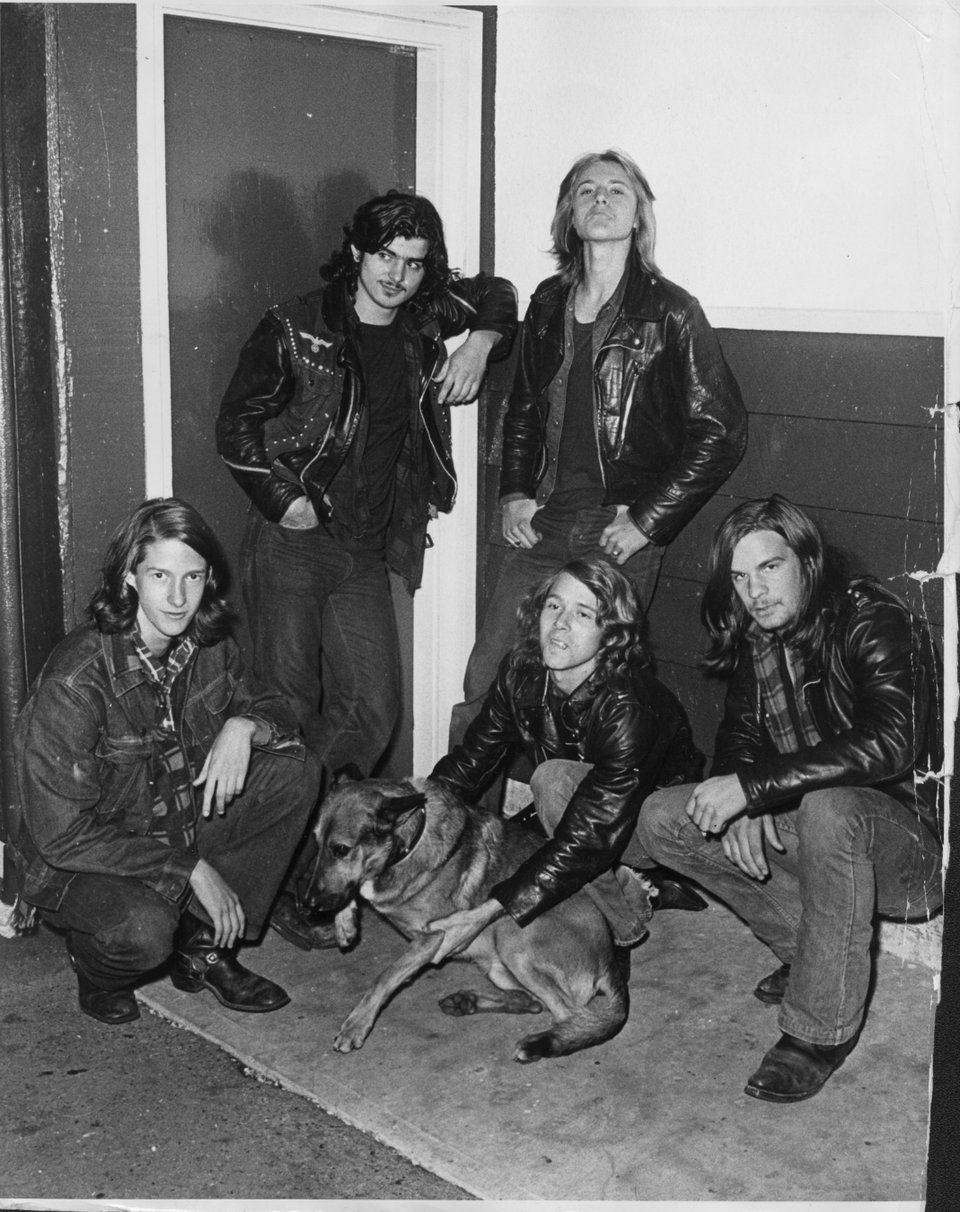 A group of five men who look to be in their 20s. They're all wearing jeans and leather jackets. One of them is holding onto a dog that looks like a German Shepherd.