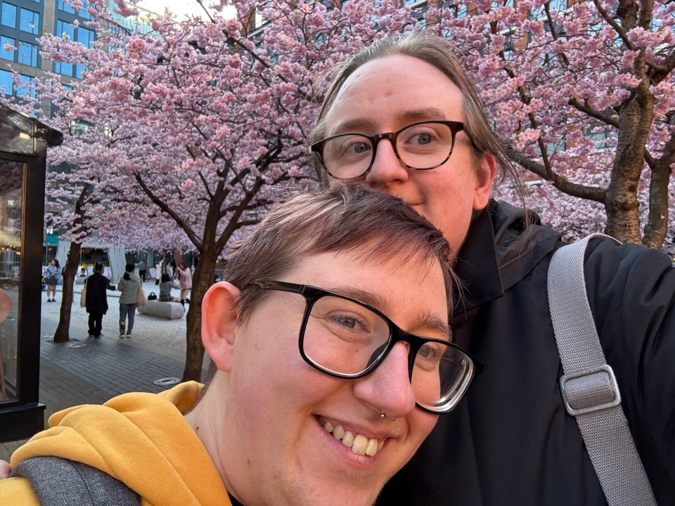 Markus and I, two white people with glasses, looking into the camera. We're stood in front of some blooming cherry blossom trees