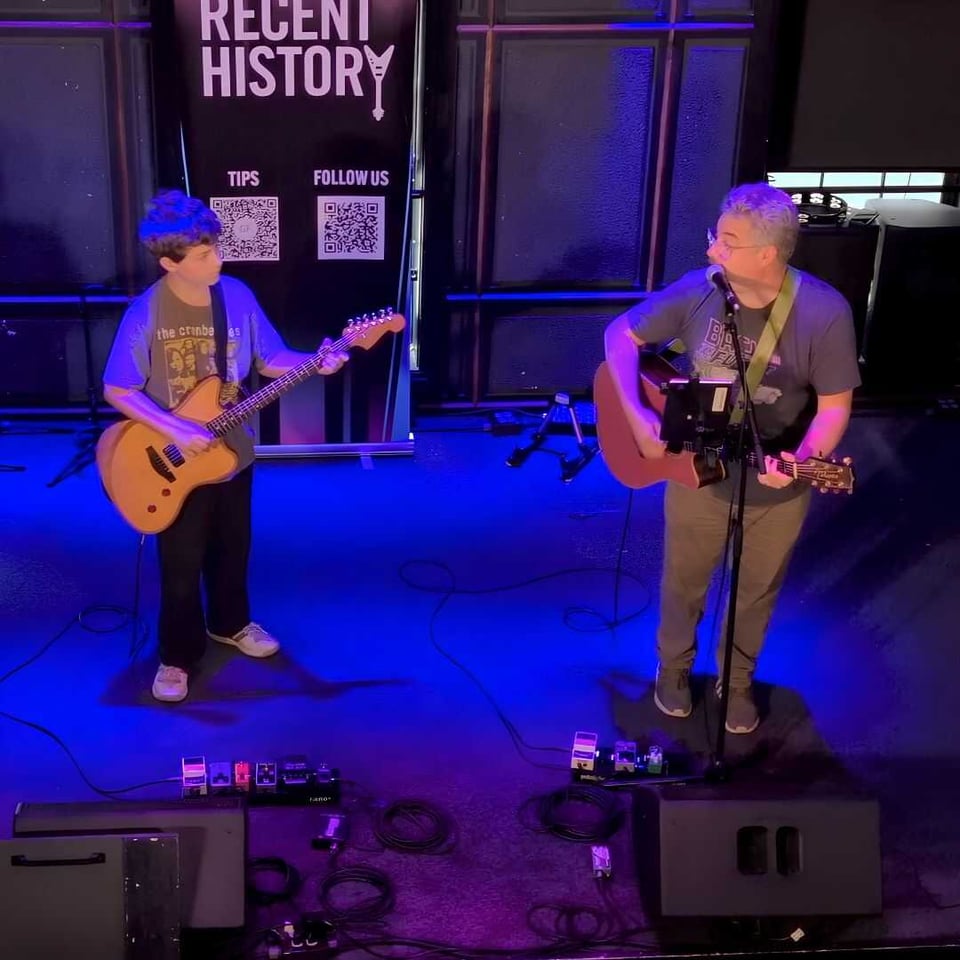 A 12-year-old boy plays electric guitar onstage next to his father, who is strumming an acoustic guitar and singing into a microphone while looking to his right at the boy.