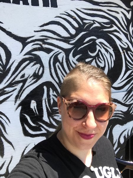 Photo of Anna, a blond person with a side-shave wearing a black t-shirt and sunglasses, takes a selfie in front of the skull mural at the Museum of Death