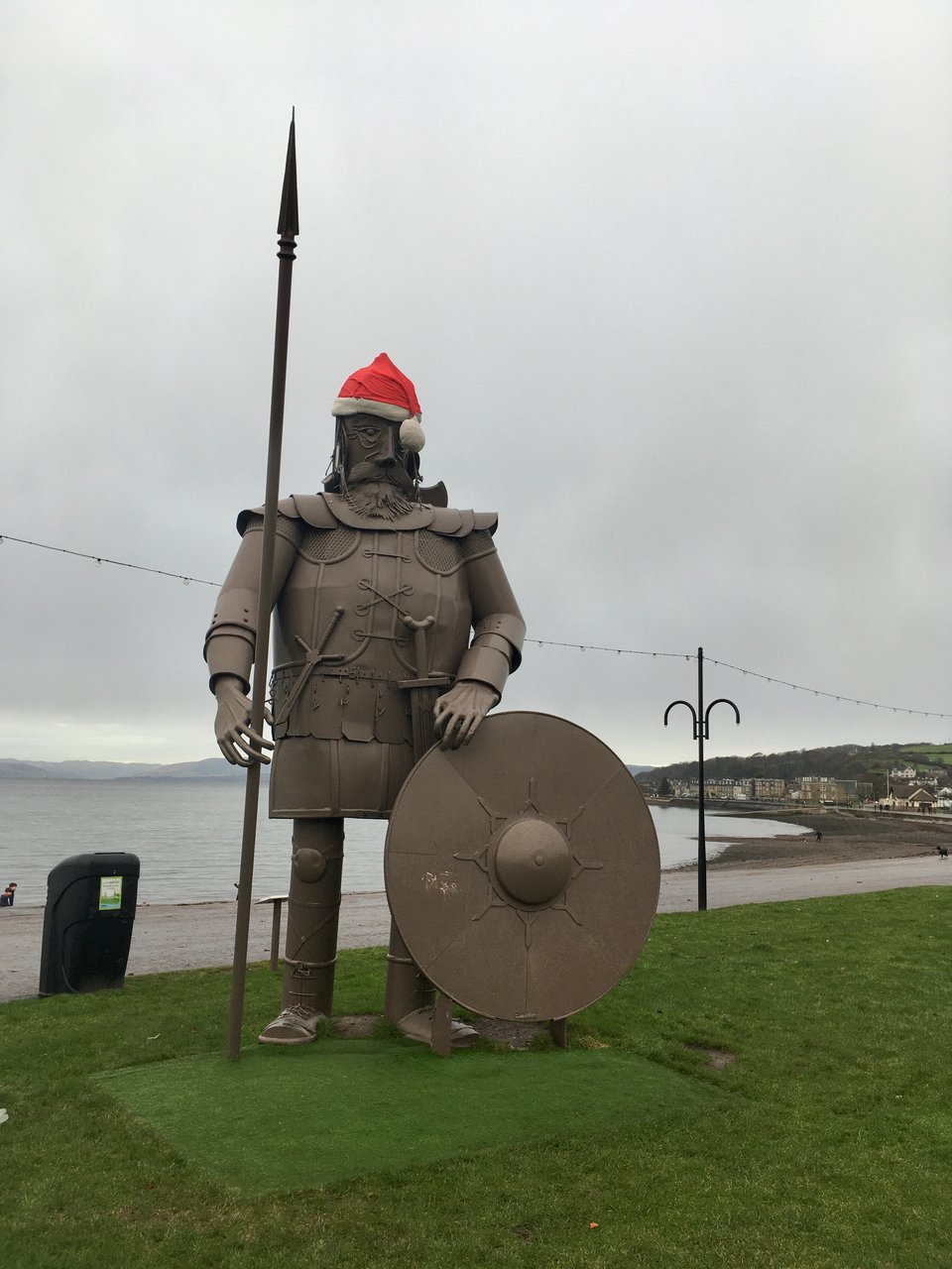 A metal statue of a Viking wears a woolly Santa hat on the promenade at Largs. The sea is pewter grey with distant, misty hills.