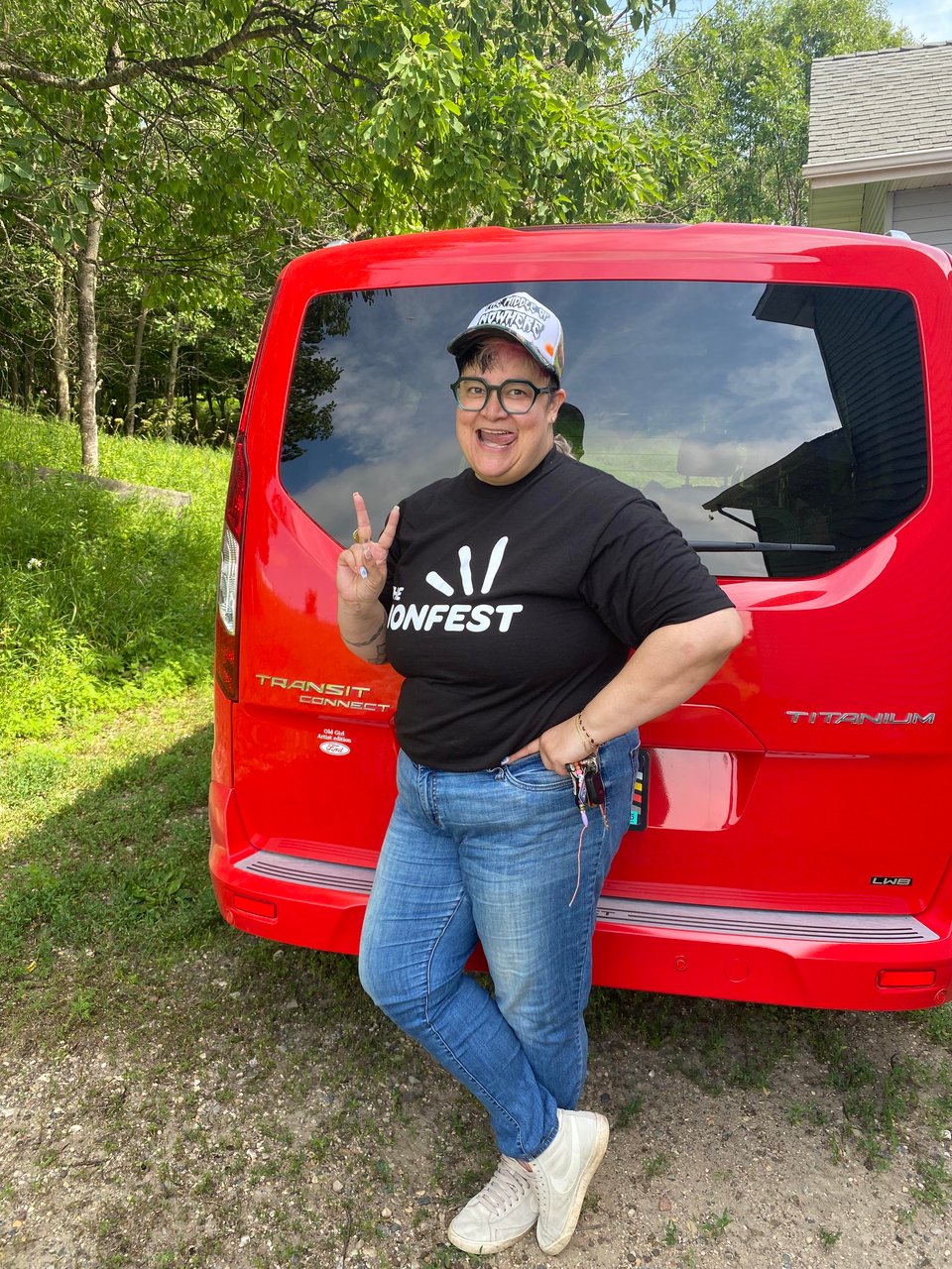 The artist standing outside in July wearing a teeshirt and jeans flashing a peace sign in front of their red van