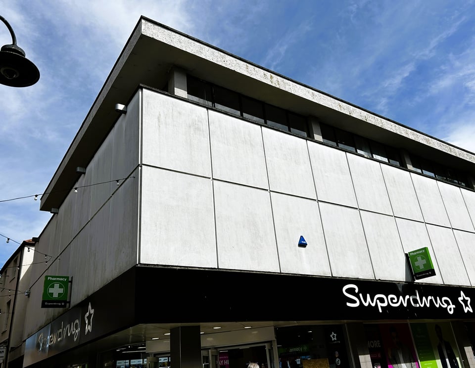 The very very plain upper storeys of a shop building. The first floor is hidden by two rows of concrete panels unadorned apart from pharmacy signs and a burgular alarm. Above this, ribbon windows are set back from the facade, beneath a concrete roof. It looks a bit like someone has lifted the lid on a shoebox.